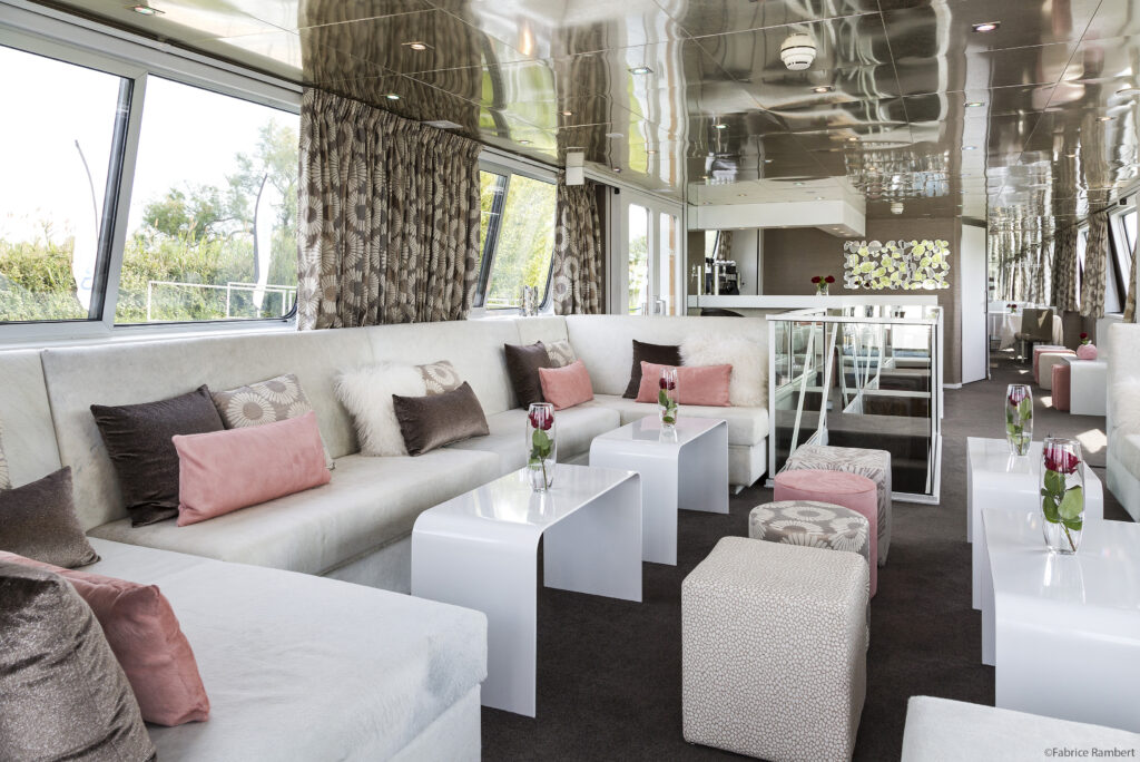 salon lounge d'une péniche croisieurope avec une banquette beige, des coussins bruns et rosés. petite tables basses blanches. Décoration signée Marine Giorgetti, architecte d'intérieur 